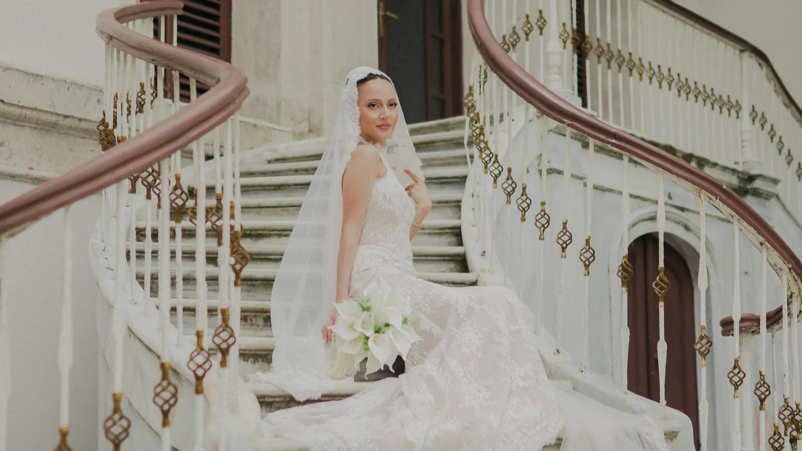 Gül seated on a curving white staircase with ornate balusters, holding white calla lilies, her lace veil falling around her, Istanbul