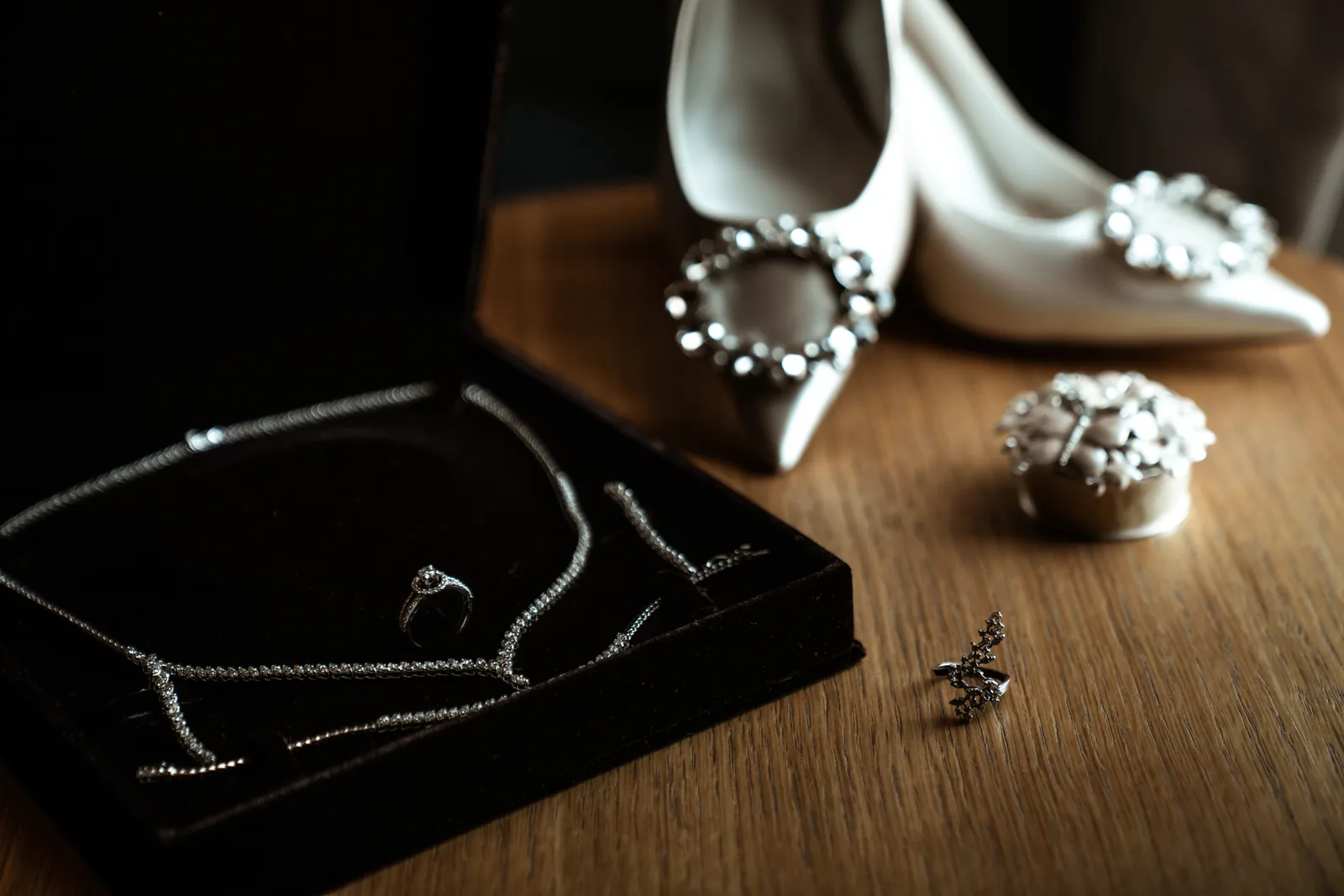 A solitaire ring and a diamond necklace in a velvet box beside satin bridal heels