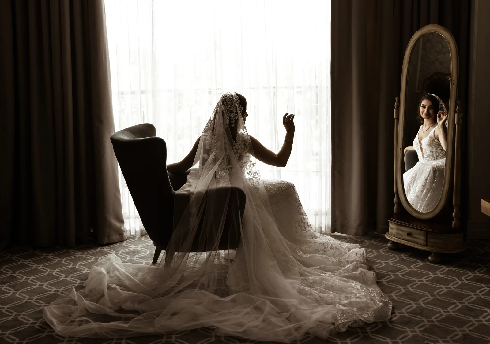 A bride in a long lace veil sits by a bright window, her face seen only in a mirror