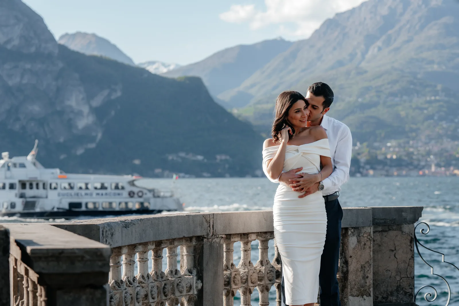 Un bacio sulla guancia a una balaustra sul lago con un traghetto alle spalle, altrove sul Lago di Como (non a Villa d'Este)