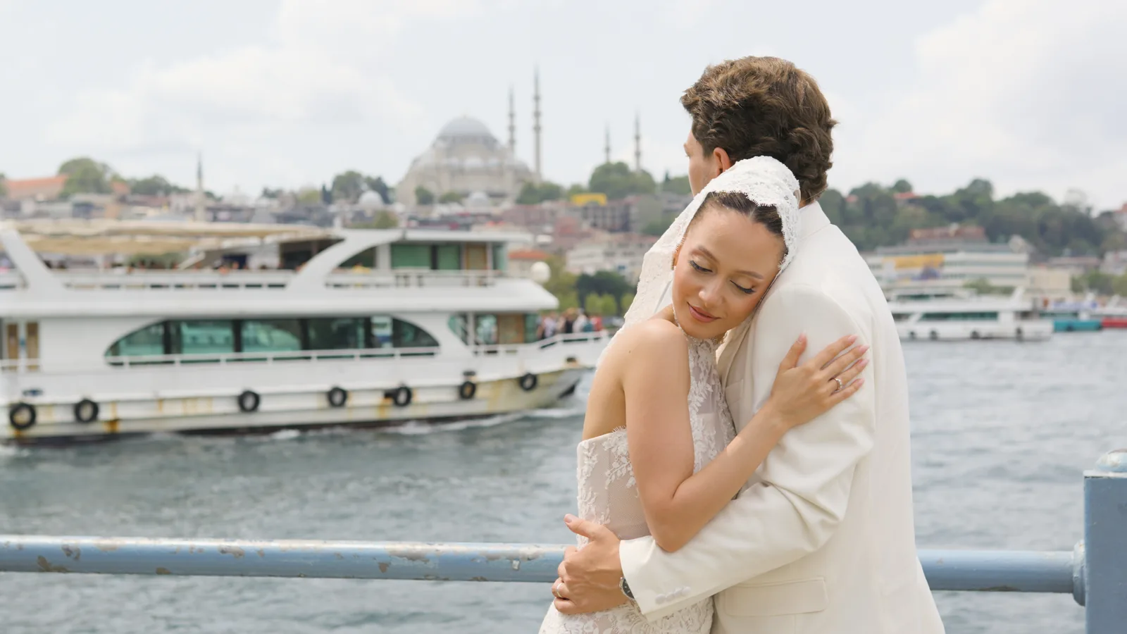 Gül and Oğuz embrace at a waterfront railing in Istanbul, a ferry and a mosque behind them