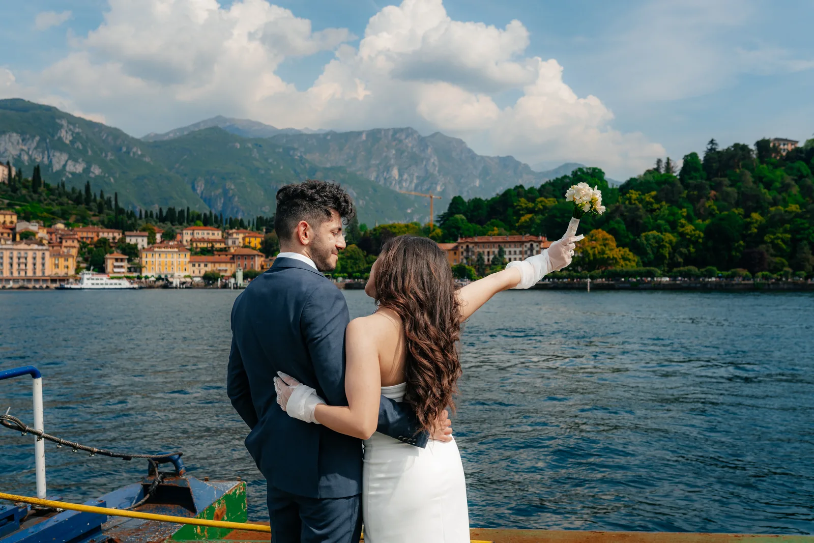 Musa and Solmaz seen from behind on the deck of a ferry on Lake Como, she raising her bouquet towards a lakeside town and the mountains, during their engagement session