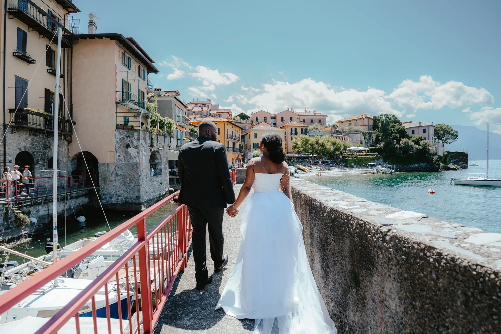 A couple seen from behind walking along a harbour wall towards pastel houses on Lake Como