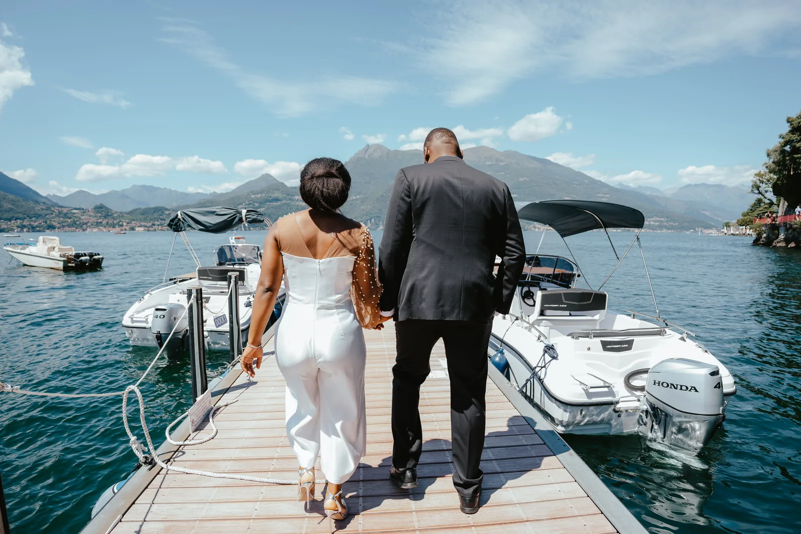 A couple seen from behind walking along a jetty between moored motorboats on Lake Como