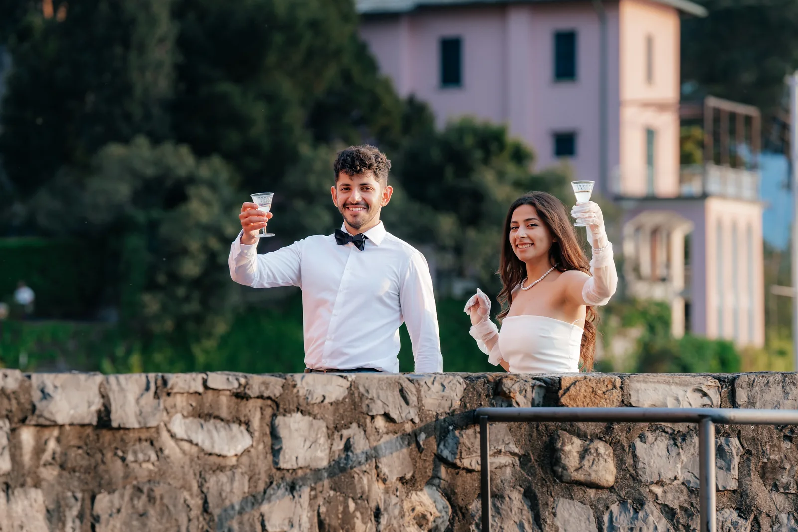 A couple raise their glasses behind a stone wall in warm evening light on Lake Como