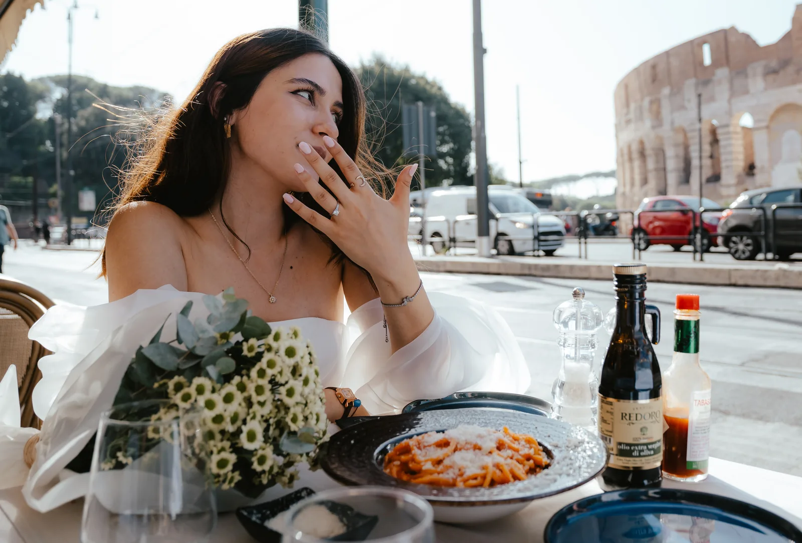 A woman in an off-the-shoulder white dress covering her mouth and showing her ring at a café table with pasta and daisies, the Colosseum across the street