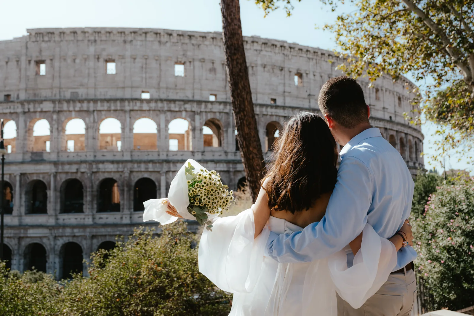 A couple seen from behind, holding each other and looking at the Colosseum in Rome