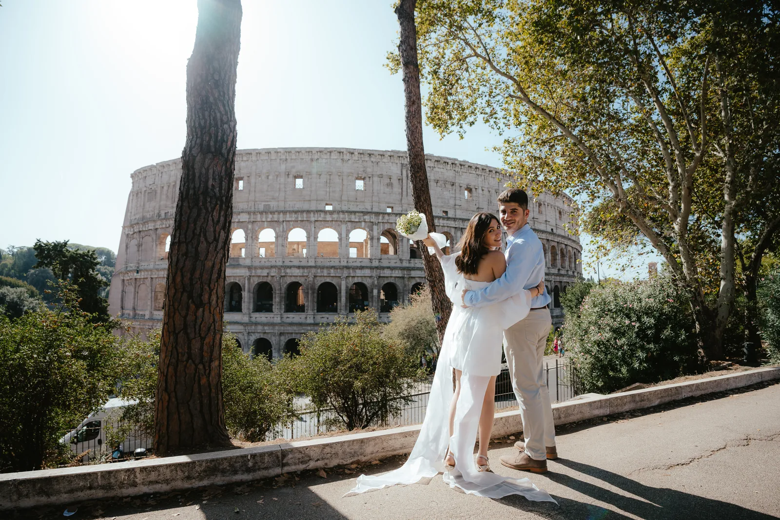 A couple embrace on a tree-lined walkway and look back at the camera, the Colosseum behind them