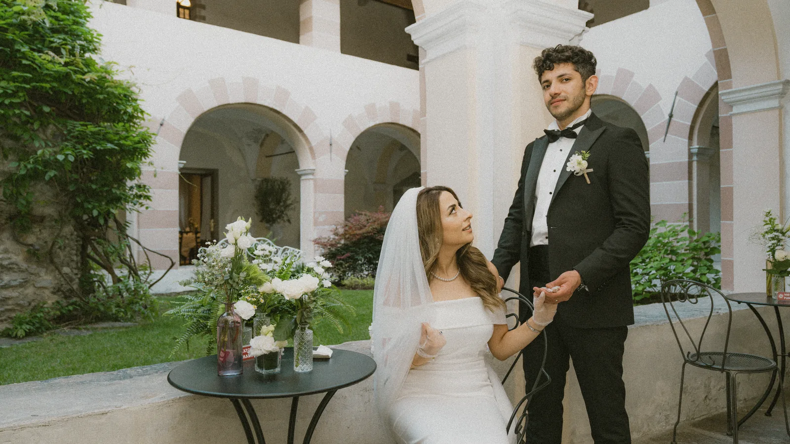 Musa and Solmaz under pink and white stone arches in Susa, Piedmont, during their pre-wedding session, she seated by a table of white flowers holding his hand