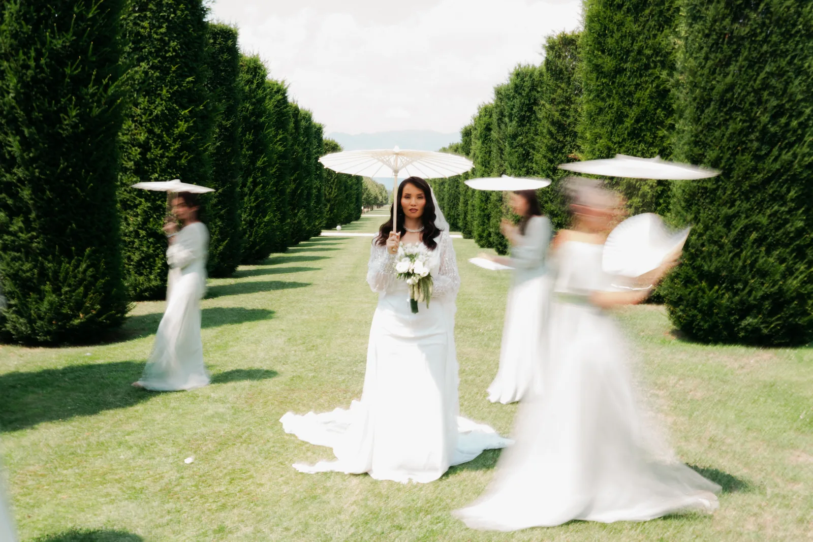 Hejrat con un parasole bianco in un viale di siepi dei giardini della Reggia di Venaria, con le damigelle alle spalle
