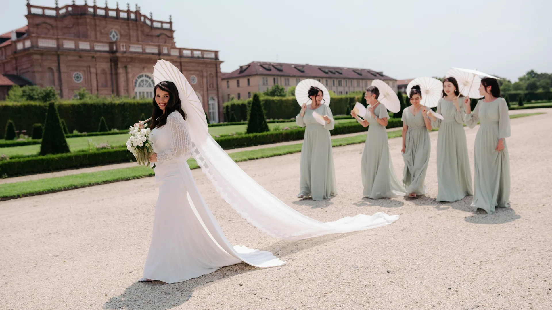 Hejrat con il bouquet e il lungo velo nei giardini della Reggia di Venaria, alle sue spalle le damigelle in verde salvia con parasoli bianchi