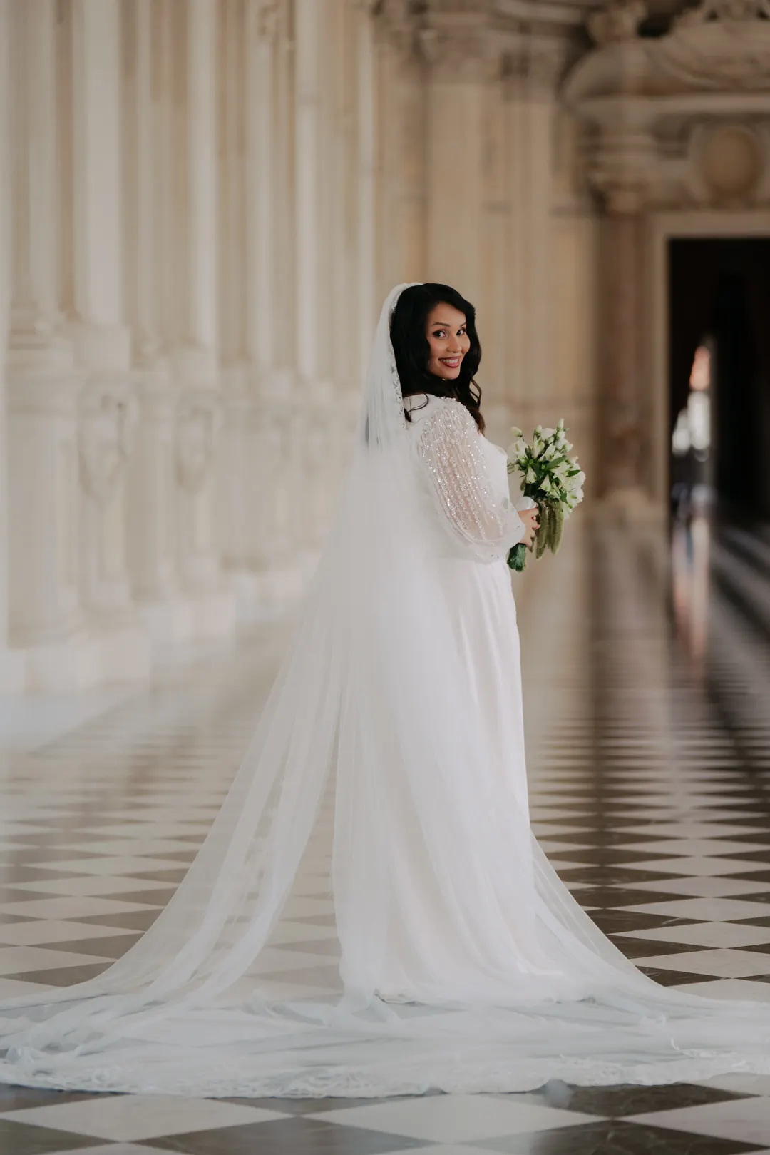 Hejrat standing alone in the long gallery of the Reggia di Venaria, its chequered floor and tall arched windows behind her, looking back over her shoulder
