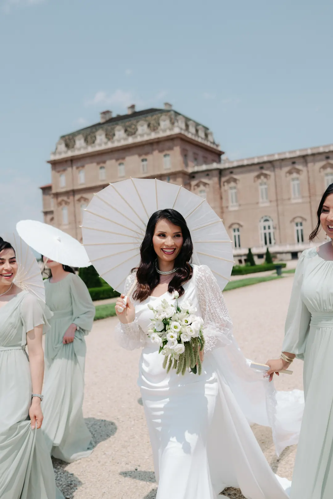 Hejrat in a white gown holding a parasol and bouquet, walking with bridesmaids in sage green dresses in front of the Reggia di Venaria Reale