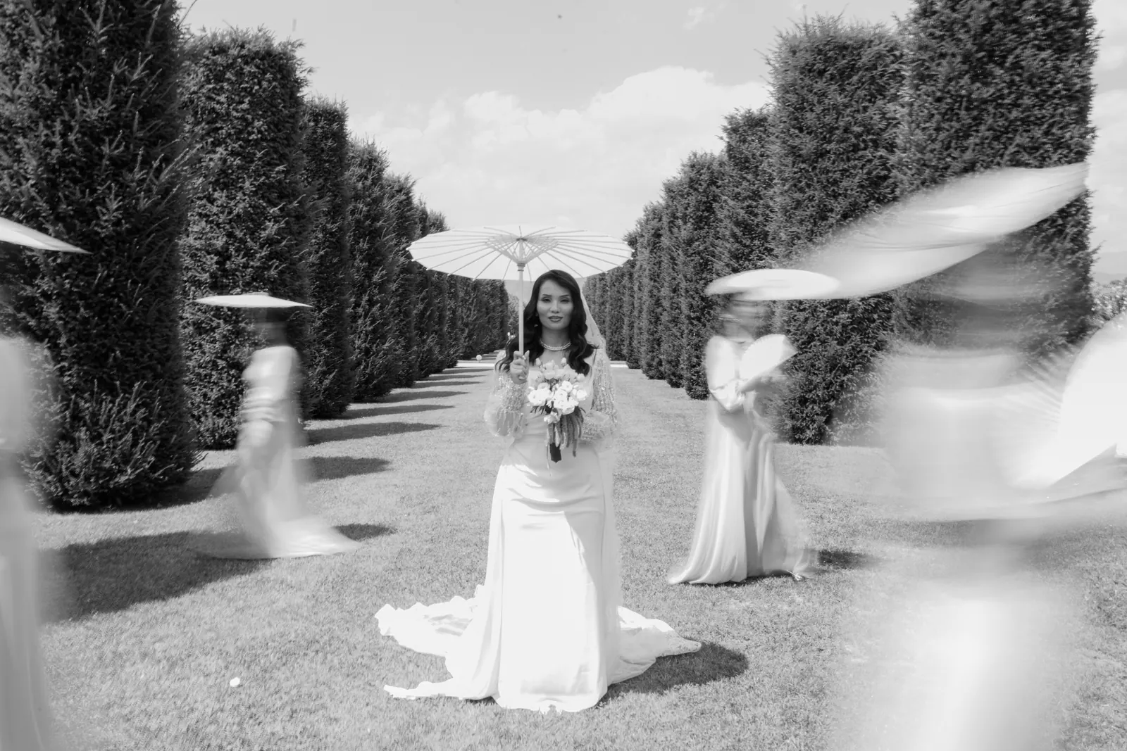 Hejrat standing still in a grass avenue between tall clipped hedges with a white parasol, bridesmaids blurred in motion around her, in black and white