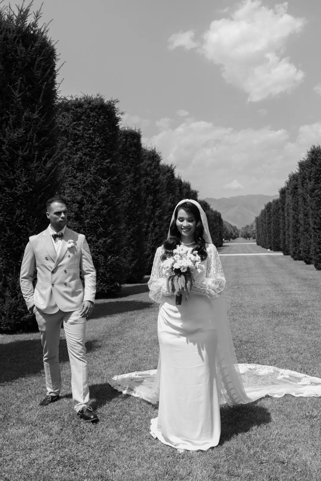 Mohammed and Hejrat standing on a long lawn between tall clipped hedges, mountains in the distance, in black and white