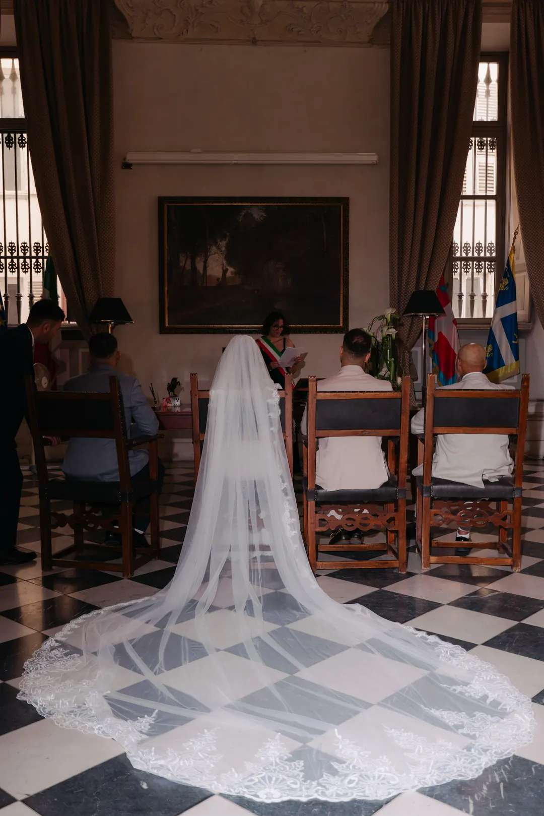 Hejrat's long lace veil spread over a chequered floor during her civil ceremony, the officiant with a tricolour sash at the desk ahead