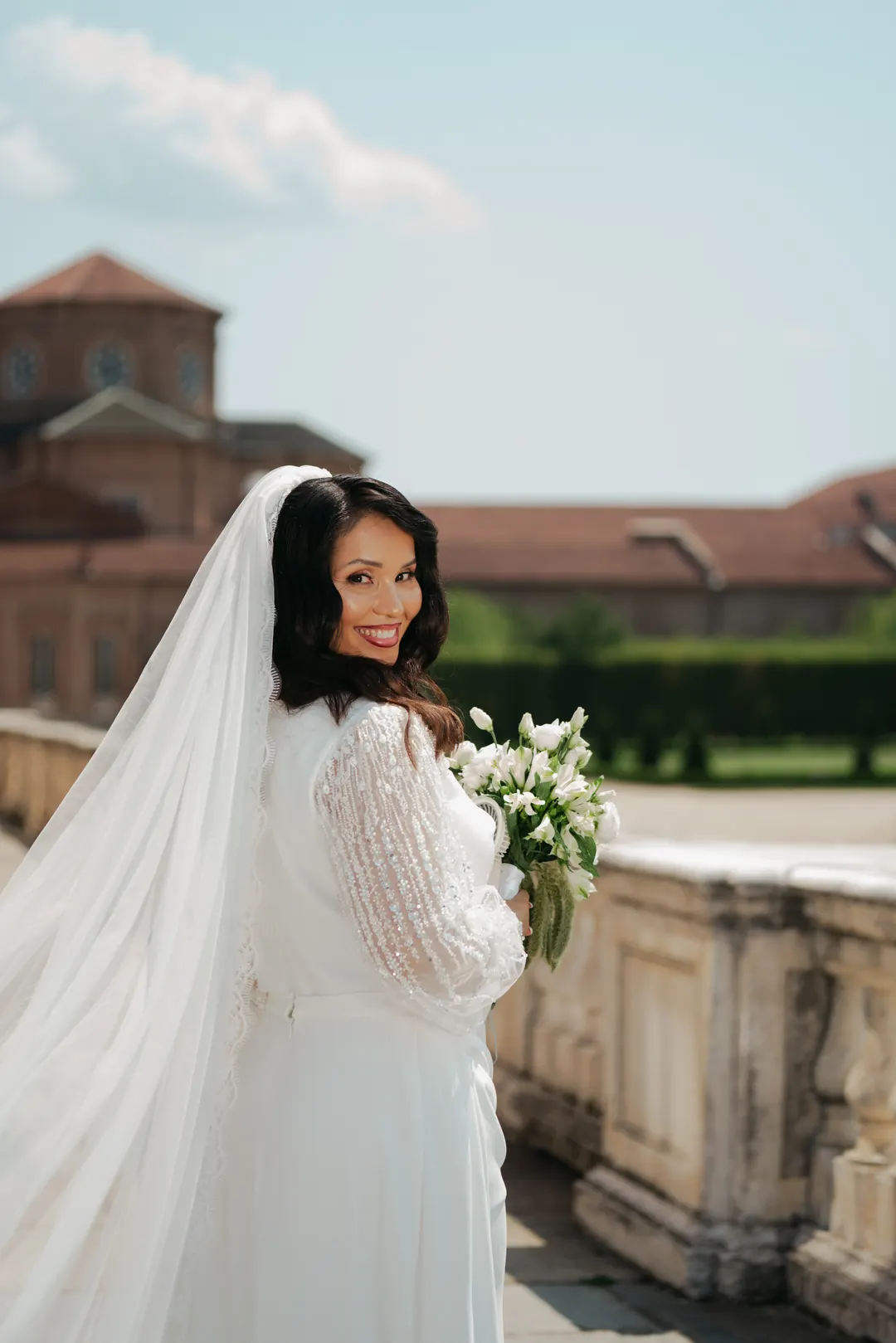Hejrat at a stone balustrade at the Reggia di Venaria, looking back over her shoulder and smiling, the palace roofs and a dome behind her