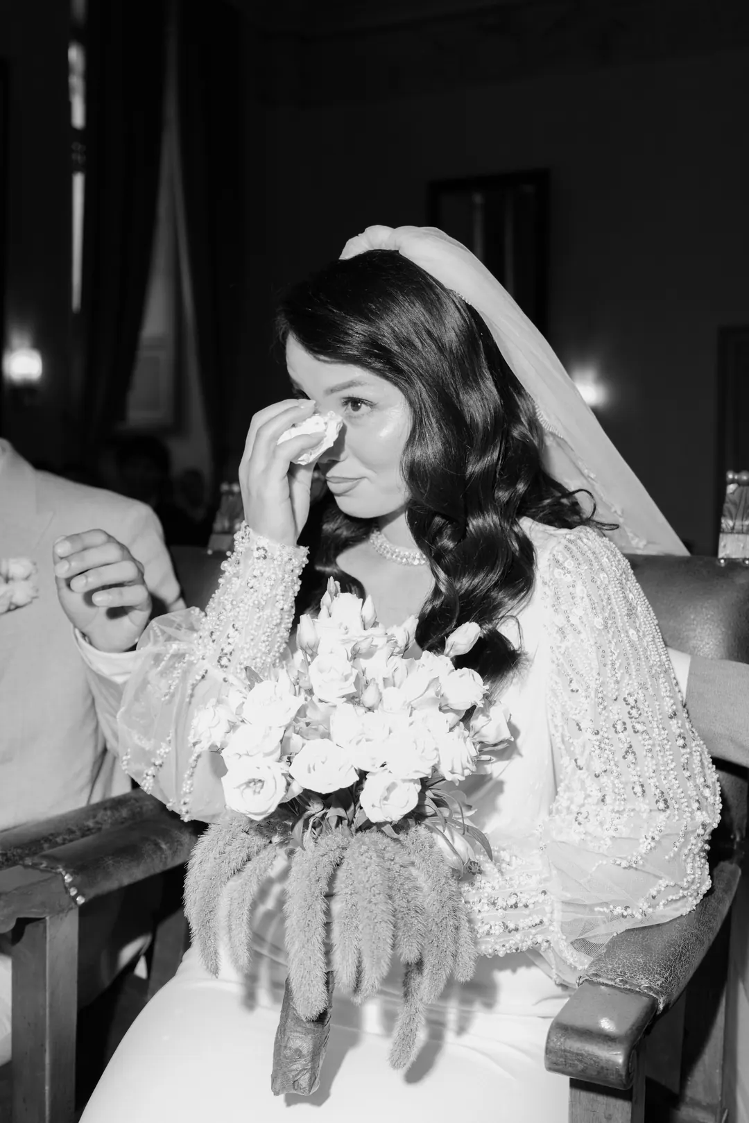 A bride wiping a tear with a tissue during her civil ceremony, holding white flowers, her veil over her hair, in black and white
