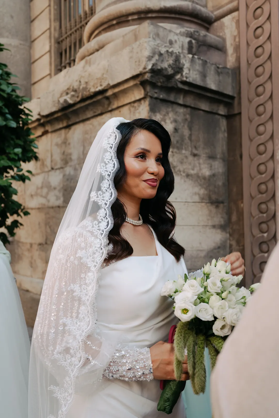 A bride in a lace-edged veil, holding white flowers with trailing amaranthus beside a carved stone column