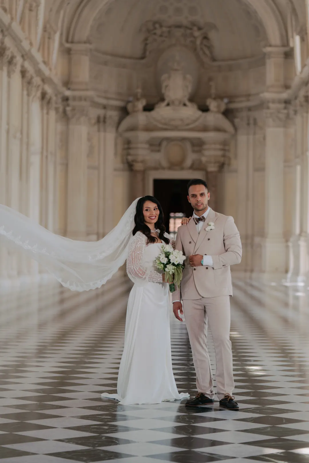 Mohammed and Hejrat standing together in a long white gallery with a chequered marble floor, her veil lifting in the air beside them