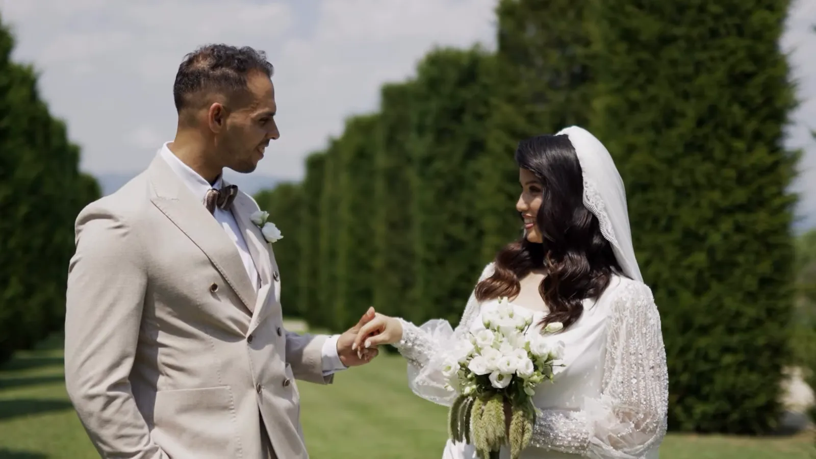 Mohammed in a beige double-breasted suit holding Hejrat's hand between tall clipped hedges, she in a beaded long-sleeved gown and veil with a white bouquet
