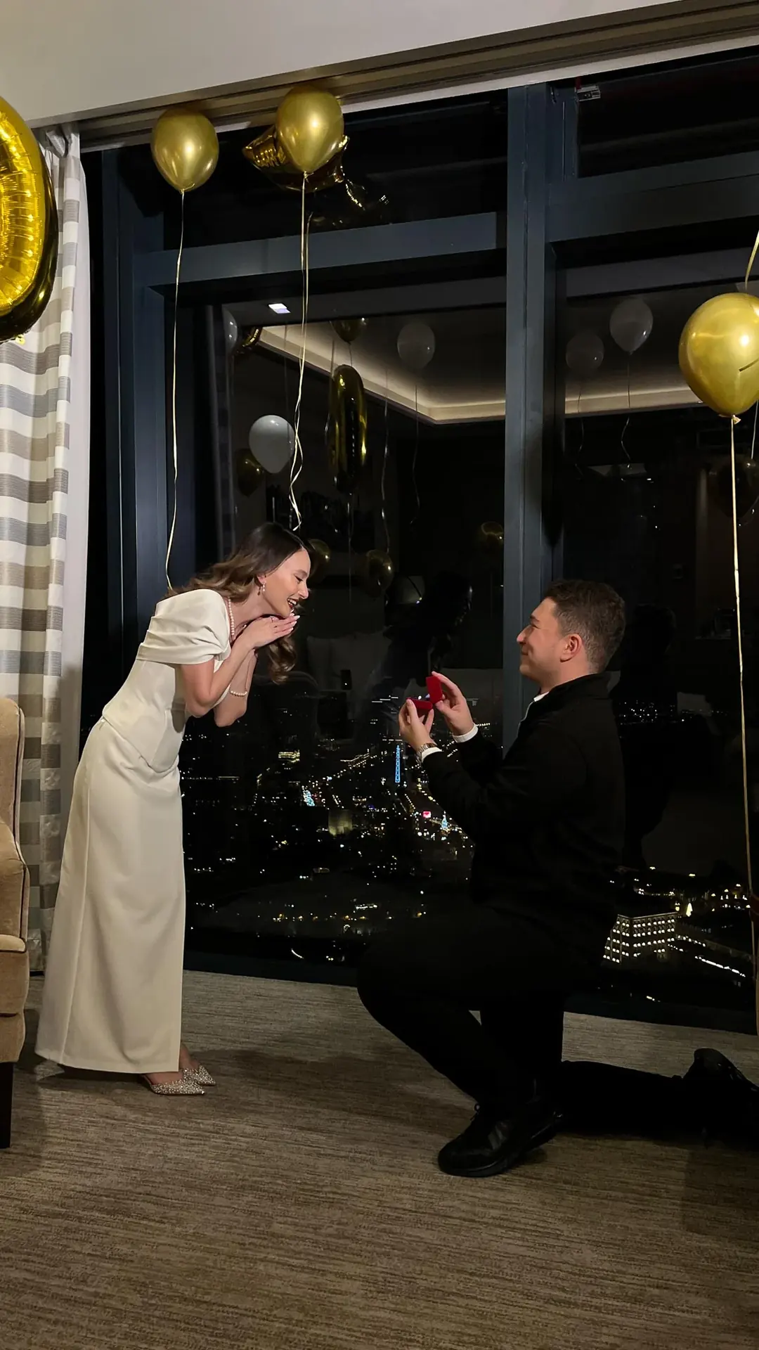 A man on one knee holding out a red ring box as a woman bends towards him with her hands at her face, in a room with gold balloons and a night city view