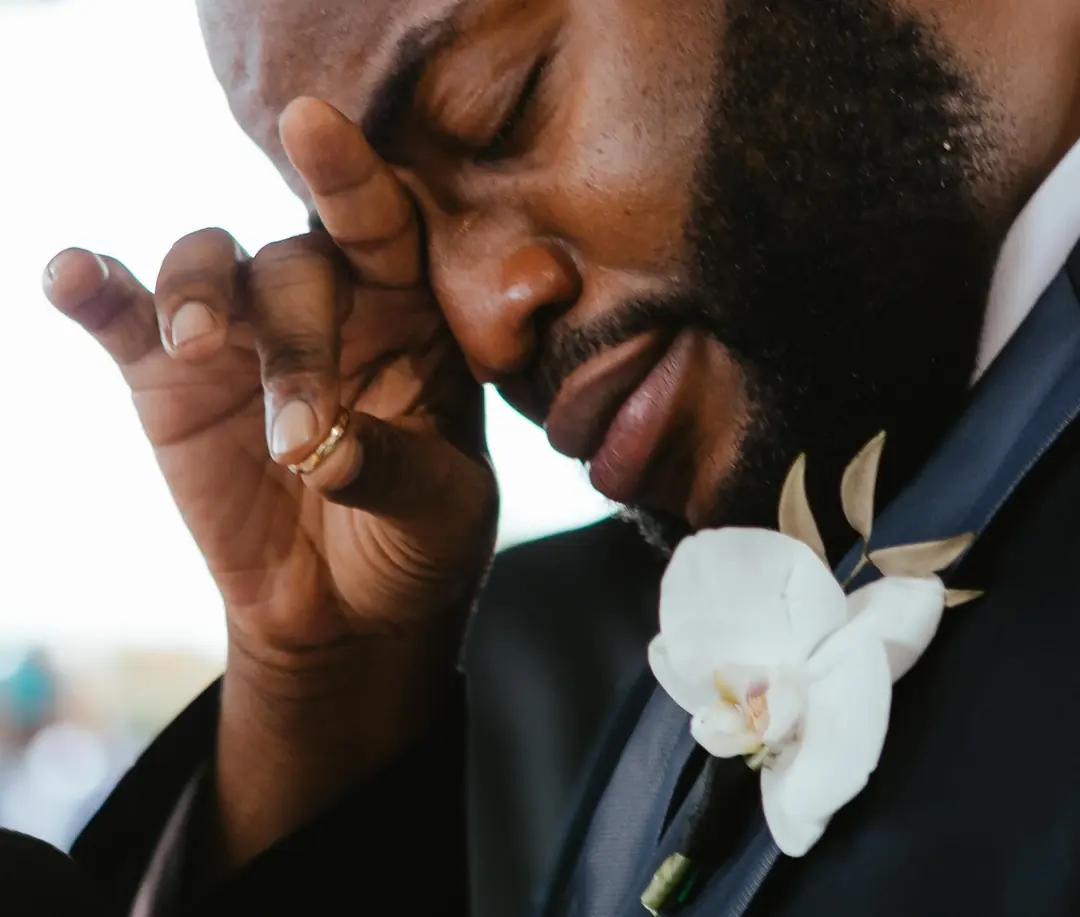 A man in a dark suit with a white orchid buttonhole wiping a tear from his eye